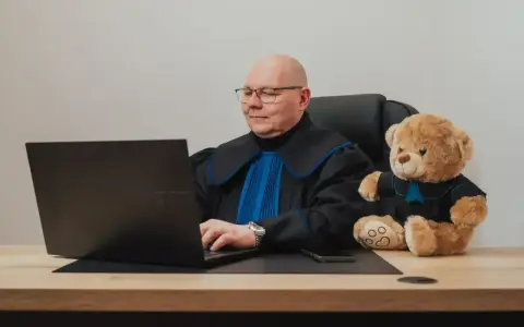 Prawnik w todze z pluszowym misiem obok, pracujący przy laptopie. Zastanawiasz się, ile kosztuje porada prawna?