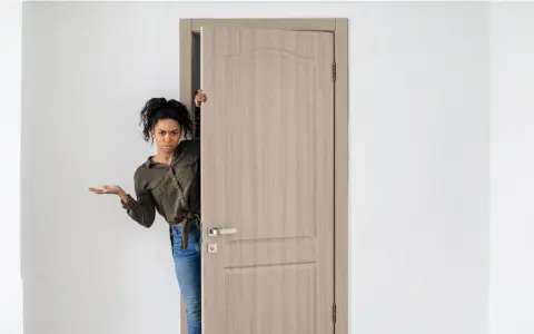Mujer asomándose por una puerta de madera clara, con expresión de duda. Los colores de puertas como esta son muy versátiles.