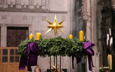 Ein Adventskranz mit vier gelben Kerzen und einem goldenen Stern, geschmückt mit lila Bändern. Seine Bedeutung liegt im Warten und Hoffen auf Weihnachten.