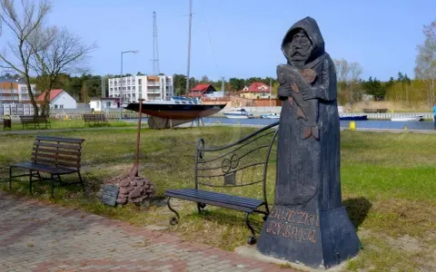 Drewniany pomnik rybaka trzymającego rybę, z napisem "Ławeczka Rybaka". W tle port z łodziami i budynkami.