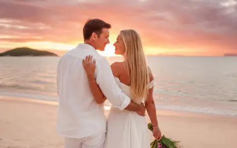 Pareja en la playa al atardecer, celebrando bodas románticas. Él, con camisa blanca, la abraza. Ella, con vestido de novia, mira al horizonte.