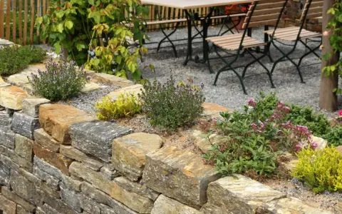 Natürliche Steinmauer mit Kräutern und Blumen, die schöne Ideen für den Garten bietet. Eine Sitzgruppe lädt zum Verweilen ein.