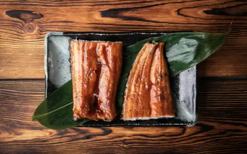 Dwa kawałki glazurowanego węgorza do sushi, podane na liściu na ciemnej zastawie, na drewnianym tle.