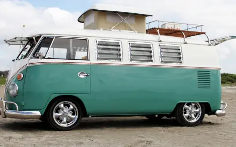 En turkos och vit volkswagen folkabuss med taklucka och takräcke står på en sandstrand.