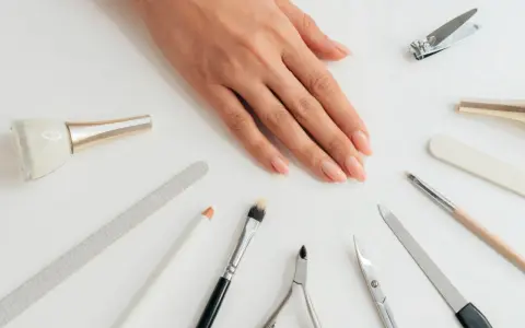 Dłoń z naturalnymi paznokciami otoczona akcesoriami do manicure. Wśród nich pilnik do paznokci naturalnych, cążki, nożyczki i pędzelki.