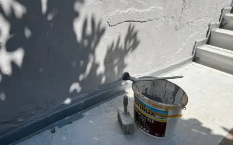Werkzeug und Farbeimer bereit zum Ausbessern des Sockelputzes. Schatten von Blättern fallen auf die Wand.