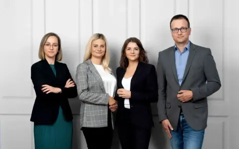 Cztery osoby w strojach biznesowych stoją obok siebie na jasnym tle.