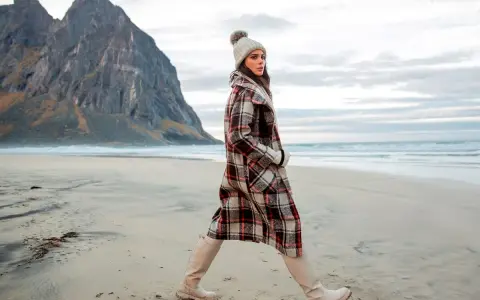 Kobieta w stylowych płaszczach w kratę damskie spaceruje po plaży, w tle góry i morze.