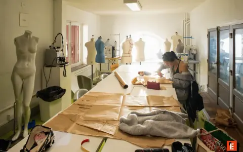 Dans un atelier d'une école d'arts, une jeune femme coupe du tissu sur une grande table. Des mannequins et du matériel de couture sont visibles.