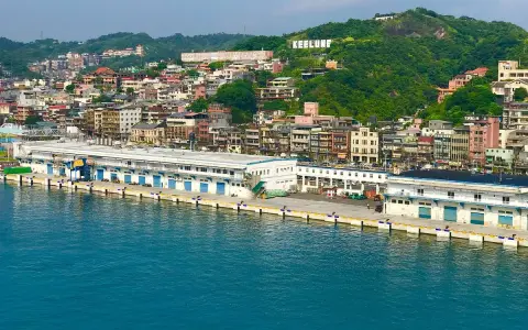 Vista panoramica del porto di Taiwan, con edifici colorati che si arrampicano sulla collina verde. La scritta "KEELUNG" domina la cima.