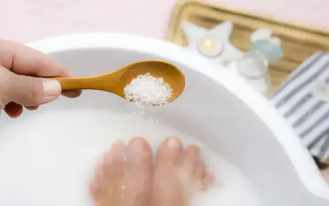 A hand holds a wooden spoon with coarse salt, about to add it to a white basin where feet are soaking. This is a relaxing foot soak with baking soda.