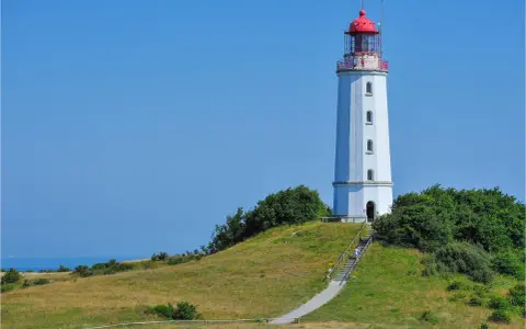Der Leuchtturm Dornbusch ragt auf einer grünen Anhöhe empor, mit dem blauen Meer im Hintergrund. Eine Treppe führt zum Eingang.