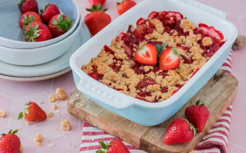 Ein frisch gebackener Erdbeer Crumble in einer hellblauen Auflaufform, garniert mit Erdbeerhälften. Frische Erdbeeren liegen daneben.