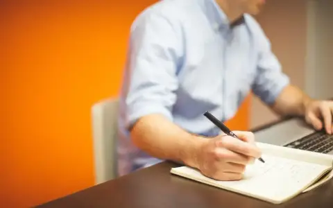 Mężczyzna w niebieskiej koszuli pisze piórem w notatniku, obok laptopa. Działa z pełną **prokura**.
