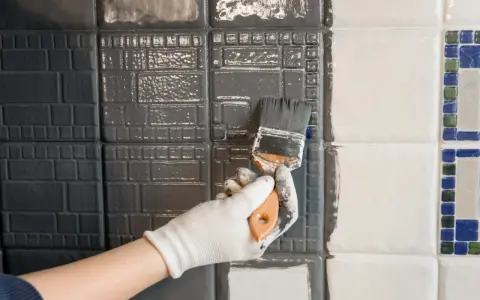 Ręka w białej rękawiczce maluje pędzlem szare płytki. Widać postęp prac, część płytek jest już pomalowana.
