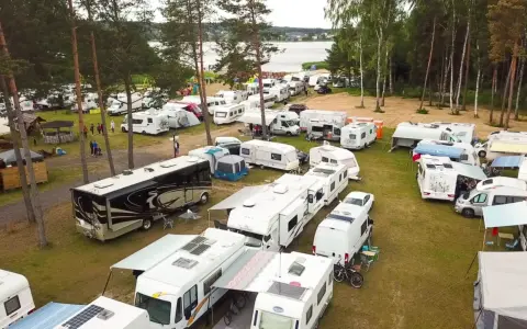 Duży zlot caravaningowy nad jeziorem. Wiele kamperów i przyczep kempingowych zaparkowanych na trawie, otoczonych drzewami.