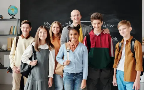 Uśmiechnięty nauczyciel i uczniowie przed tablicą. Czy szkoła społeczna to miejsce, gdzie tak dobrze się czują?