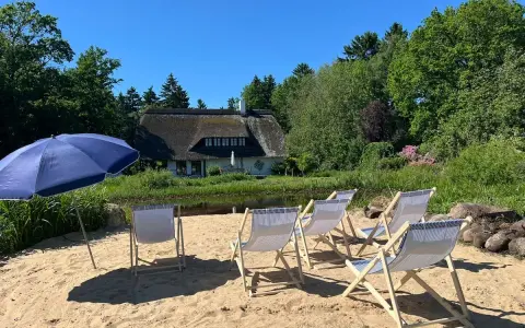 Sonnige Tage am Stedener See: Liegestühle und Sonnenschirm warten auf Besucher vor einem Reetdachhaus.