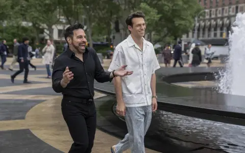 Il cast di And Just Like That, Mario Cantone e Billy Eichner passeggiano ridendo vicino a una fontana in una giornata nuvolosa.