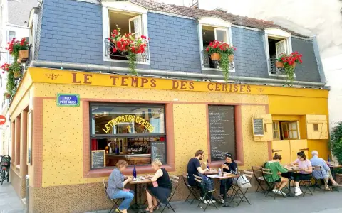 Café "Le Temps des Cerises" con un encantador estilo francés, balcones floridos y gente disfrutando en sus mesas exteriores.