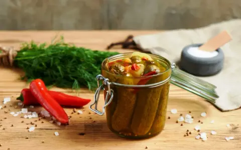 Słoik pełen pikantnych ogórków po meksykańsku, obok świeży koperek i czerwone papryczki chili.