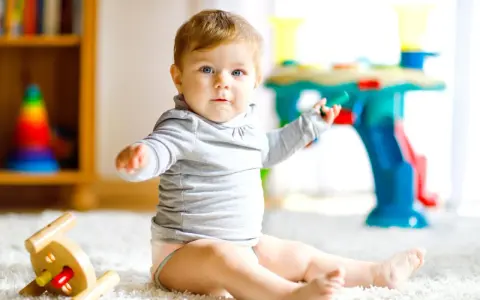 Ein neugieriges 8 Monate altes Baby mit blauen Augen sitzt auf einem flauschigen Teppich und spielt mit Spielzeug.