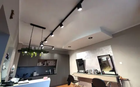 Nowoczesne oświetlenie w salonie fryzjerskim tworzy przytulną atmosferę. Reflektory na szynach i wisząca lampa z zielenią podkreślają styl wnętrza.