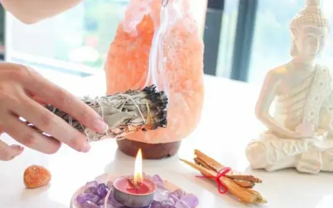 Manos sostienen un manojo de salvia humeante, junto a una lámpara de sal rosa, una vela encendida y una estatua de Buda. Esencial para la limpieza energética de casa.