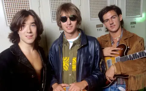 Les membres de Talk Talk posent devant des casiers. Le guitariste tient une guitare électrique.