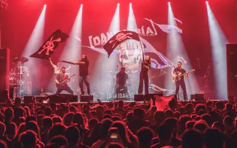 Una banda toca en un escenario iluminado con luces rojas, ondeando banderas piratas. El público, con trece roeis, anima la actuación.