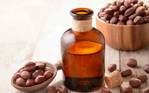 Butelka z bursztynowego szkła z płynnym złotym olejkiem jojoba na włosy, obok drewnianej łyżki z orzechami jojoba.