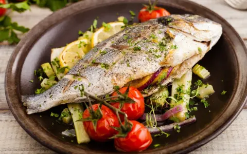 Poisson entier, probablement une truite au barbecue, servi avec des tomates cerises, des quartiers de citron et des herbes fraîches sur une assiette marron.