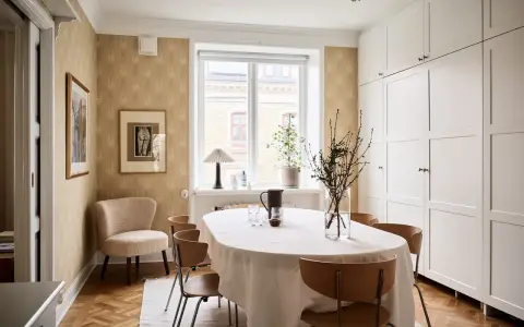 Comedor con mesa cubierta, sillas de madera y pared con papel tapiz dorado. Armarios blancos y suelo de parqué.