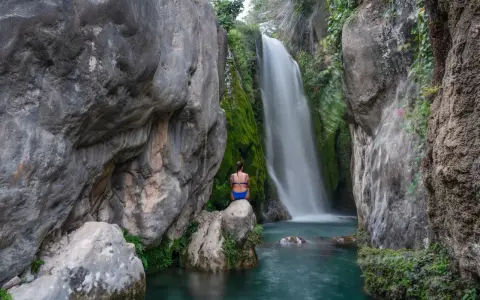 Una mujer sentada en una roca contempla la cascada de las fuentes del Algar, rodeada de vegetación y rocas.