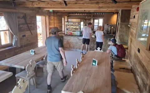 Wnętrze schroniska na Hali Kondratowej. Drewniane stoły i ławy, bar z ekspozycją ciast i napojów. Kilka osób odpoczywa po wędrówce.