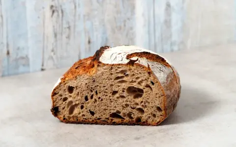 Ein halbes, knuspriges schnelles Sauerteigbrot mit großer Krume und rustikaler Kruste liegt auf einem hellen Untergrund vor einer hellblauen Holzvertäfelung.
