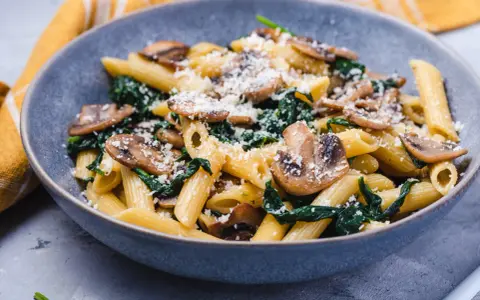 Penne z pieczarkami i szpinakiem, posypane parmezanem, w szarej misce.
