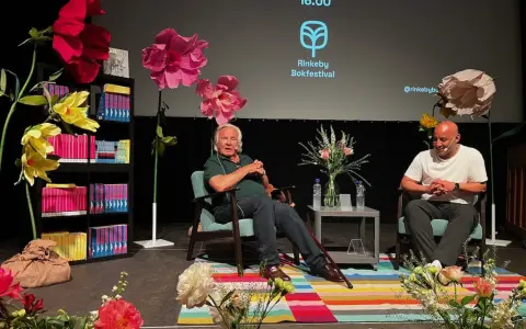 Jan Guillou delar fakta på Rinkeby Bokfestival. Scenen är dekorerad med stora blommor och bokhyllor.