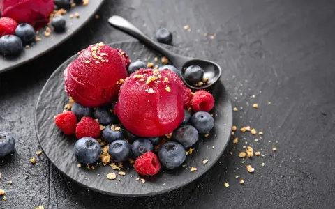 Zwei Kugeln Campari Sorbet mit frischen Beeren und Streuseln auf einem schwarzen Teller.