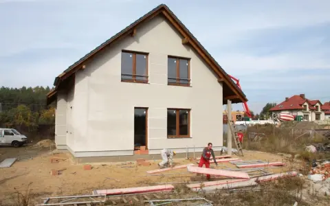 Budowa domu w toku. Pracownicy układają płyty, a w tle widać betonowóz. Czas budowy zależy od wielu czynników.