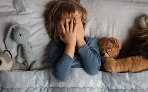 Kind versteckt Gesicht, umgeben von Kuscheltieren. Die Angst um das Kind ist spürbar, es sucht Trost bei seinen Freunden.