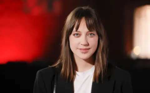 Maria Sobocińska z niebieskimi oczami i grzywką, ubrana w czarną marynarkę i białą koszulkę, uśmiecha się lekko na tle czerwonego światła.