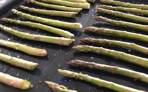 Färska sparrisar ligger på en ugnsplåt, redo att rostas i ugnen. Saltkorn glittrar på de gröna stjälkarna.