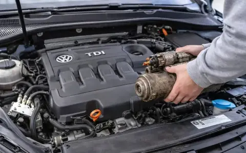 Ręce trzymają zużyty rozrusznik, gdy samochód nie chce odpalić. Widoczny silnik TDI Volkswagena.