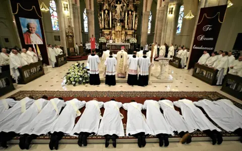 Klerycy w białych szatach leżą na posadzce kościoła, w geście pokory. Obok nich stoją inni duchowni, a w tle widać ołtarz i portret papieża.
