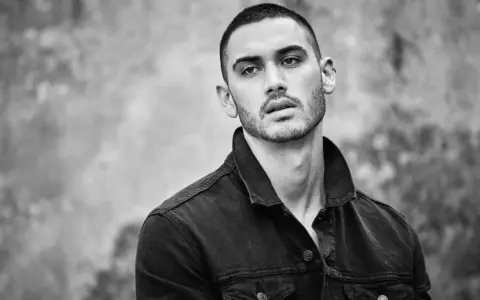 Hombre joven con un corte de pelo buzz cut, viste chaqueta vaquera oscura. Mirada intensa.