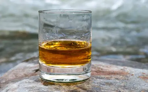 Ein Glas goldener Whisky steht auf einem Stein am Flussufer. Die ideale trinktemperatur für diesen edlen Tropfen.