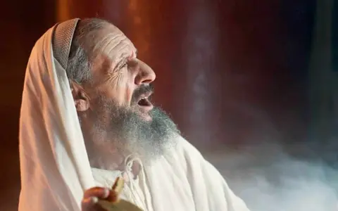 An elderly man, possibly Zechariah in the Bible, with a grey beard and wearing a white robe and head covering, cries out with his mouth open.