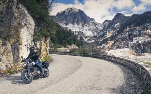 Un motociclista percorre una strada tortuosa tra le montagne della Toscana, con cave di marmo sullo sfondo.