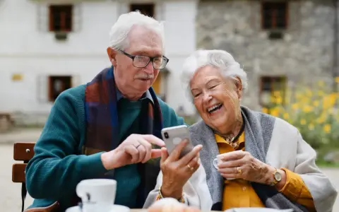 Uśmiechnięta para seniorów korzysta z telefonu, odkrywając nowe aplikacje dla seniorów. Radość i wspólne chwile przy kawie.